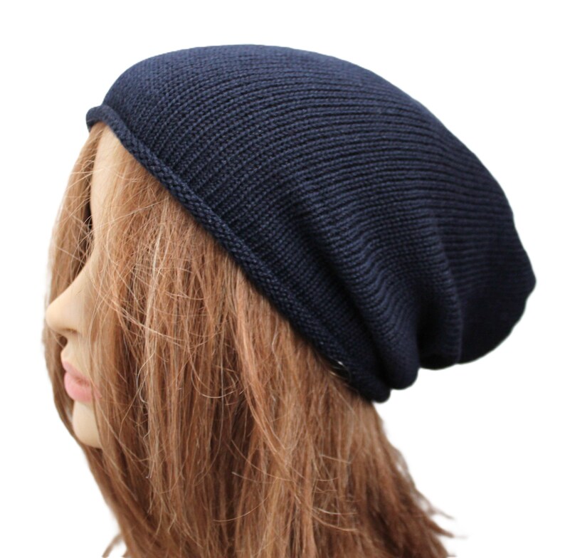 May include: A dark navy blue beanie hat with a fine knit texture and a simple rolled edge hem. The hat features a slouchy silhouette that sits comfortably on the head, providing a clean and uniform appearance with its consistent dark blue color.
