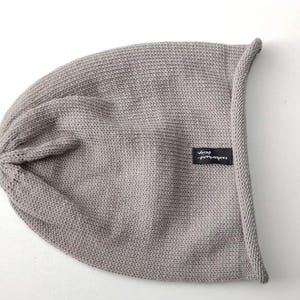 May include: A light gray knit beanie with a rolled edge and a small black rectangular fabric tag featuring the white text zauberhand design. The hat is constructed from a fine, uniform knit fabric with a gathered top and a soft, slouchy shape.