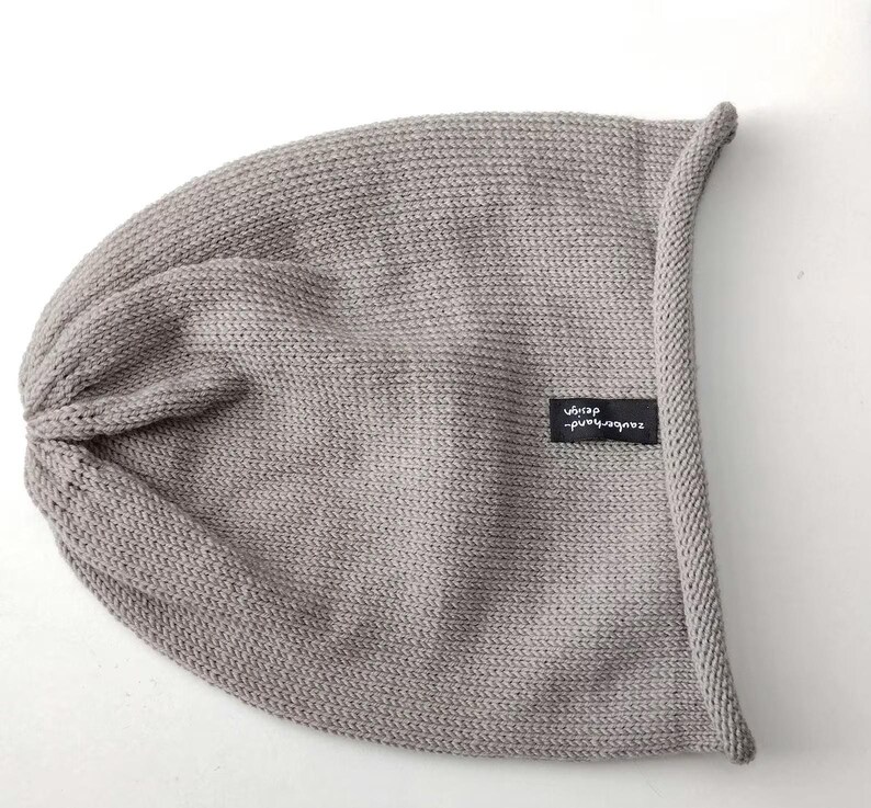May include: A light gray knit beanie with a rolled edge and a small black rectangular fabric tag featuring the white text zauberhand design. The hat is constructed from a fine, uniform knit fabric with a gathered top and a soft, slouchy shape.