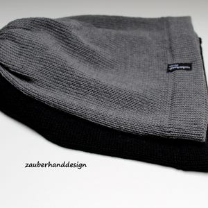 May include: Two knit beanies stacked together, one in charcoal gray and one in black. Each beanie features a textured knit pattern and a small black fabric tag on the side with the white text zauberhanddesign. The text zauberhanddesign is also printed in black on the surface below the beanies.