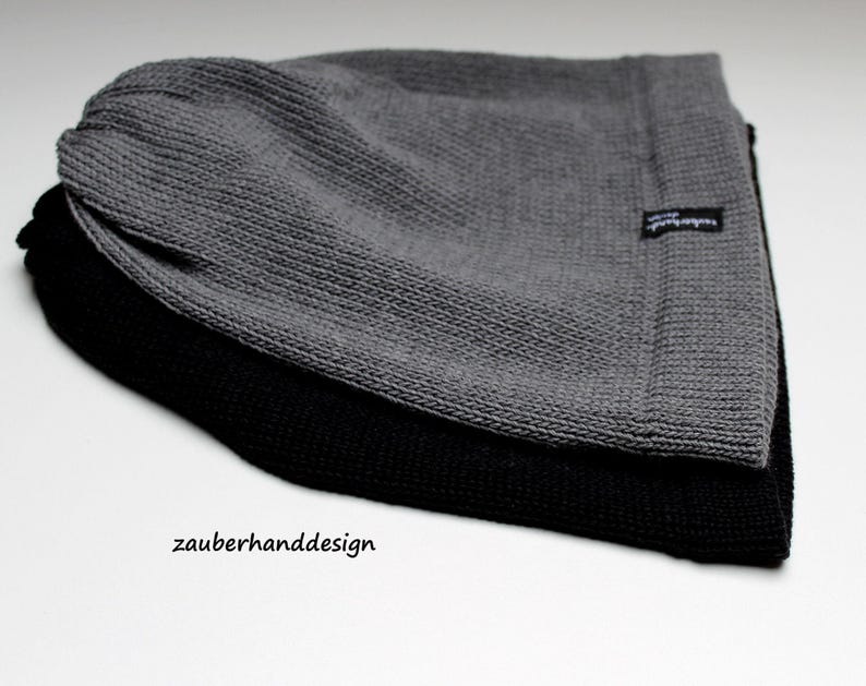 May include: Two knit beanies stacked together, one in charcoal gray and one in black. Each beanie features a textured knit pattern and a small black fabric tag on the side with the white text zauberhanddesign. The text zauberhanddesign is also printed in black on the surface below the beanies.