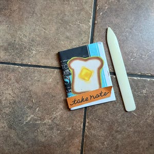 Peut inclure: Un petit livret orné d'une illustration de tranche de pain grillé avec une noisette de beurre, une bande de papier orange portant le texte manuscrit take note, et un plioir en os blanc cassé placé à côté du livret sur une surface carrelée.