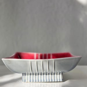 May include: A gray ceramic bowl with a rectangular shape and a red interior. The bowl has a raised base with vertical lines and a flared rim. The bowl is likely vintage or retro.