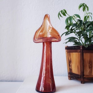 May include: A red and amber glass vase with a flared top and a slender neck. The vase is sitting on top of two books, one titled "The Smithsonian Experience" and the other titled "Mother Goose".