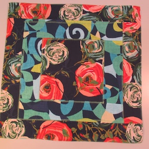 May include: A square fabric placemat with a dark blue background and a colorful floral pattern. The pattern features pink, red, green, and blue flowers with swirls and geometric shapes.