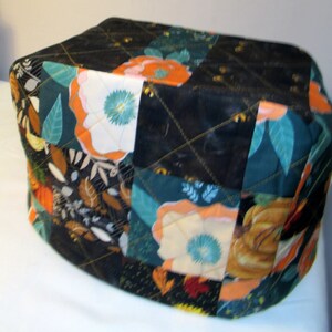 May include: A quilted fabric cover with a patchwork design featuring floral and geometric patterns in shades of black, teal, orange, and white.