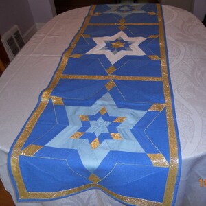 May include: A blue table runner with star patterns in shades of blue, white, and gold. The runner has a gold border and is placed on a white tablecloth. The star design is a geometric pattern.
