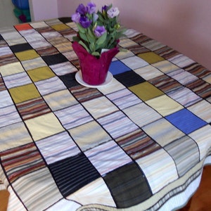 May include: A patchwork tablecloth with a grid pattern made from various shirt fabrics. The squares feature stripes, solids, and textures in colors like blue, black, brown, and yellow. A purple flower pot with flowers sits on the table.