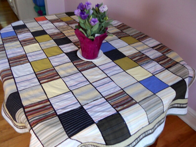 Peut inclure: Une nappe patchwork avec un motif quadrill&eacute; fait de diff&eacute;rents tissus de chemise. Les carr&eacute;s pr&eacute;sentent des rayures, des unis et des textures dans des couleurs comme le bleu, le noir, le marron et le jaune. Un pot de fleurs violet avec des fleurs est pos&eacute; sur la table.