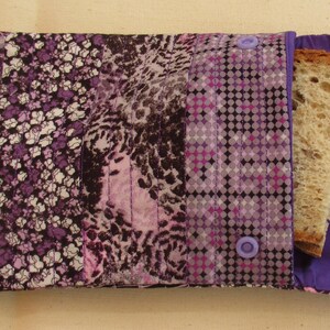 May include: A rectangular fabric sandwich bag featuring a patchwork design of purple, black, and white floral, animal, and geometric patterns. The pouch is secured with two light purple plastic snap buttons and contains a slice of whole grain bread.