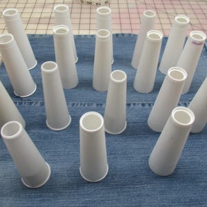 May include: A collection of fifteen empty white plastic thread cones arranged in a grid pattern. Each cone features a tapered cylindrical shape with a hollow center and a slightly wider base, designed for holding spools of thread for sewing machines.