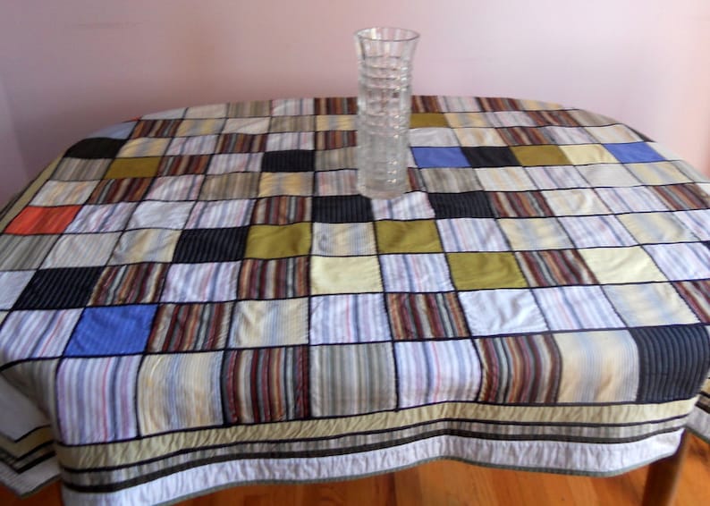 Peut inclure: Une nappe ovale &agrave; motifs avec un design patchwork aux couleurs vari&eacute;es, dont le bleu, le noir et le jaune. Un vase en verre transparent textur&eacute; est pos&eacute; sur la table. La nappe pr&eacute;sente un motif &agrave; rayures.