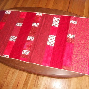 May include: A rectangular red table runner with a variety of red fabrics and patterns. The runner features vertical stripes of solid red, patterned red, and red with white floral accents. A thin white border frames the edges.