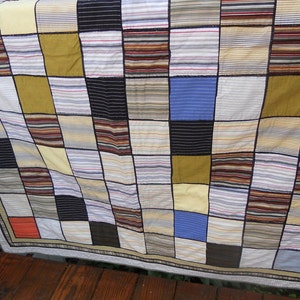 Peut inclure: Une courtepointe patchwork avec un motif quadrill&eacute;, pr&eacute;sentant des carr&eacute;s de diff&eacute;rentes couleurs et tissus ray&eacute;s. La courtepointe comprend des nuances de jaune, de bleu, de marron et de noir, avec une bordure neutre. La courtepointe est drap&eacute;e sur une surface en bois.