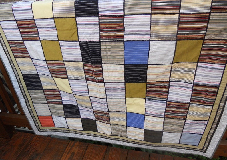 Peut inclure: Une courtepointe patchwork avec un motif quadrill&eacute;, pr&eacute;sentant des carr&eacute;s de diff&eacute;rentes couleurs et tissus ray&eacute;s. La courtepointe comprend des nuances de jaune, de bleu, de marron et de noir, avec une bordure neutre. La courtepointe est drap&eacute;e sur une surface en bois.