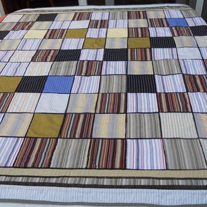 Peut inclure: Une courtepointe patchwork avec un motif quadrill&eacute;, compos&eacute;e de carr&eacute;s de diff&eacute;rents tissus. Les carr&eacute;s comprennent des tissus ray&eacute;s et unis dans des couleurs comme le bleu, le noir, le jaune et le marron. La courtepointe a une bordure ray&eacute;e.