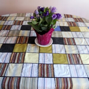 Peut inclure: Une nappe patchwork color&eacute;e avec des carr&eacute;s de tissus ray&eacute;s et unis bleus, jaunes, noirs et marrons. Un pot de fleurs violettes est pos&eacute; sur la nappe. La nappe mesure environ 150 cm de diam&egrave;tre.