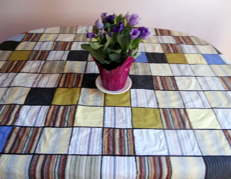 Peut inclure: Une nappe patchwork color&eacute;e avec des carr&eacute;s de tissus ray&eacute;s et unis bleus, jaunes, noirs et marrons. Un pot de fleurs violettes est pos&eacute; sur la nappe. La nappe mesure environ 150 cm de diam&egrave;tre.