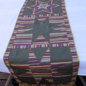 May include: A decorative table runner featuring a star pattern. The runner has a central design of large, dark green stars and triangles, with a striped background in shades of purple, green, and gold. The edges are trimmed with gold fringe.