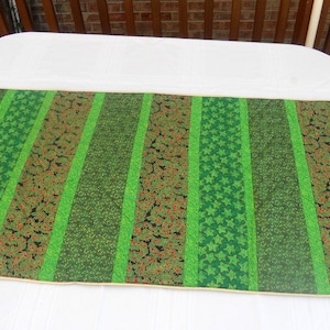 May include: A green and brown table runner with a striped pattern. The runner features alternating stripes of solid green, green star patterns, and brown floral patterns. The edges are trimmed with a gold border. The runner is placed on a white surface.