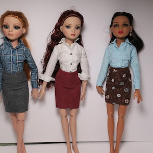 May include: Three dolls wearing different outfits. The doll on the left is wearing a blue plaid shirt and a gray skirt. The doll in the middle is wearing a white shirt with black polka dots and a burgundy skirt. The doll on the right is wearing a light blue shirt and a brown skirt with a floral pattern.