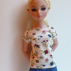 May include: A doll with blonde hair wearing a white t-shirt with a floral print and blue jeans.