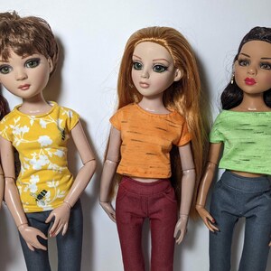 May include: Four dolls wearing different colored tops and jeans. The dolls have long hair and are wearing different colored tops. The tops are all short-sleeved and have a casual style.