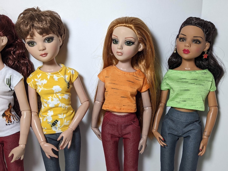 May include: Four dolls wearing different colored tops and jeans. The dolls have long hair and are wearing different colored tops. The tops are all short-sleeved and have a casual style.
