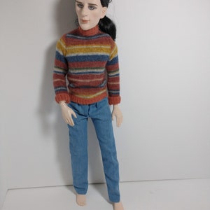 May include: A doll wearing a striped sweater with blue, red, yellow, and brown stripes. The doll is wearing blue jeans and has black hair.