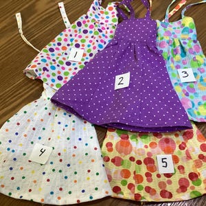 May include: Five colorful, handmade doll dresses. The dresses feature polka dots and floral patterns in various colors, including purple, white, yellow, and blue. Each dress has a unique design and numbered tag.