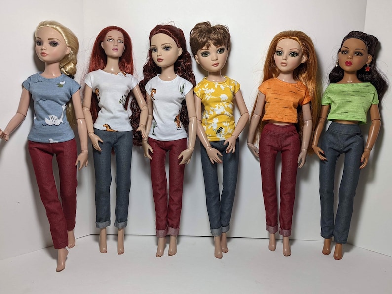 May include: Six dolls wearing different colored tops and jeans. The tops are all short-sleeved and have different patterns. The jeans are all blue and have a cuffed hem.