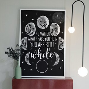 May include: Black and white print with a quote that reads "No matter what phase you're in you are still whole". The quote is surrounded by moon phases with floral designs.