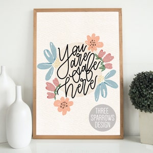May include: A framed art print with the words "You are safe here" in black script, surrounded by watercolor flowers in shades of blue, pink, and peach. The print has a light beige background and a wooden frame. The words "THREE SPARROWS DESIGN" are at the bottom.