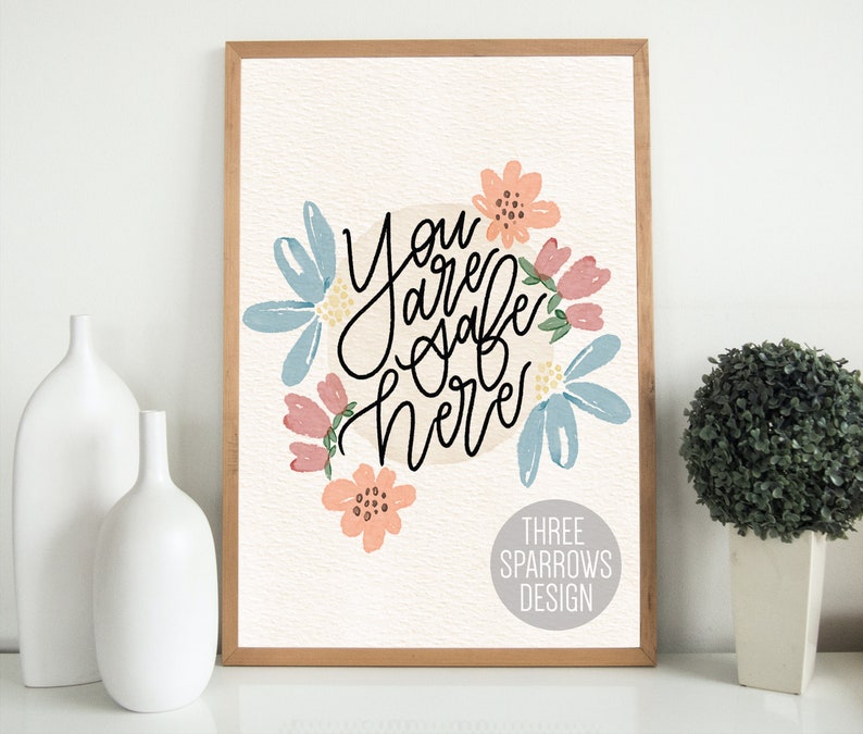 May include: A framed art print with the words "You are safe here" in black script, surrounded by watercolor flowers in shades of blue, pink, and peach. The print has a light beige background and a wooden frame. The words "THREE SPARROWS DESIGN" are at the bottom.