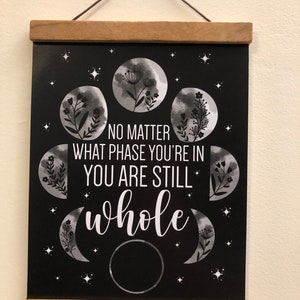 May include: Black and white chalkboard sign with a quote about phases of the moon. The quote reads "No matter what phase you're in you are still whole". The sign is decorated with images of the moon in different phases and flowers.