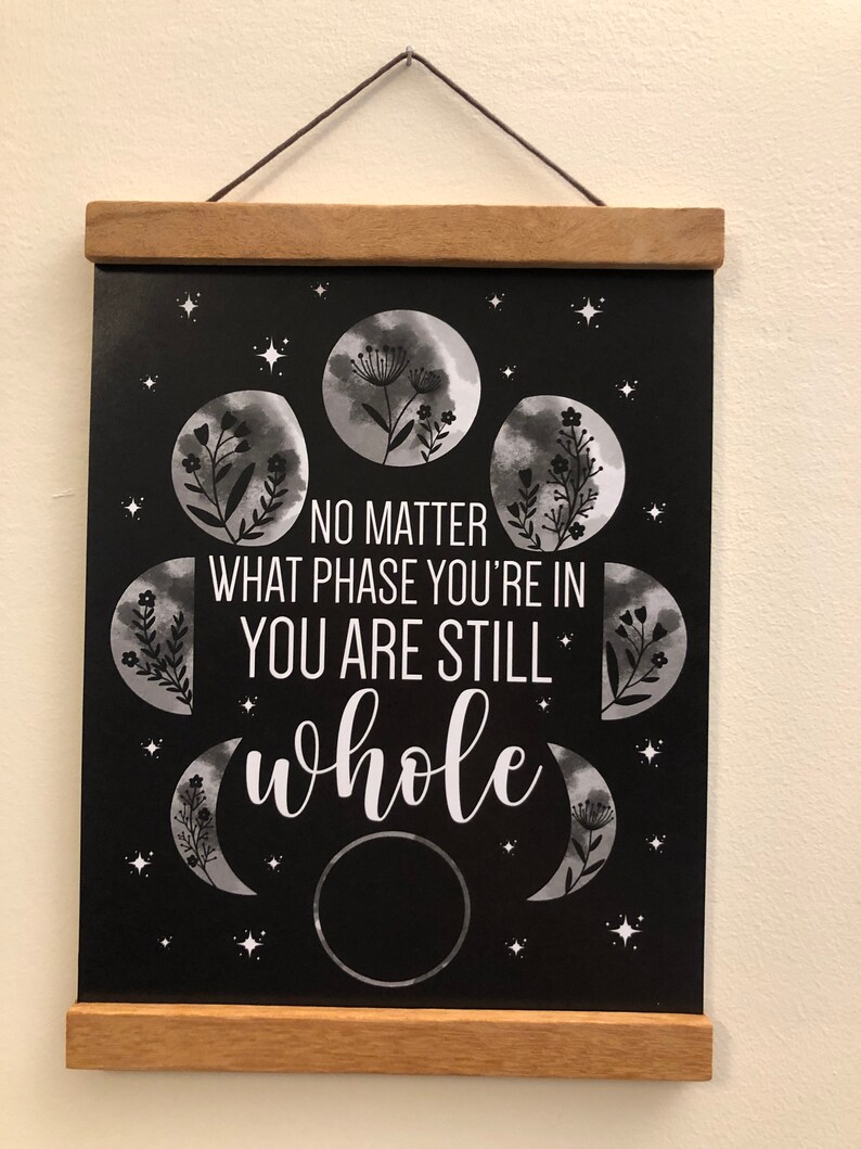 May include: Black and white chalkboard sign with a quote about phases of the moon. The quote reads "No matter what phase you're in you are still whole". The sign is decorated with images of the moon in different phases and flowers.