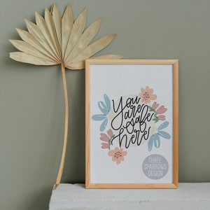 May include: A framed print with the words "You are safe here" in black script, surrounded by pastel-colored floral illustrations. The frame is light wood. A dried palm leaf is to the left.