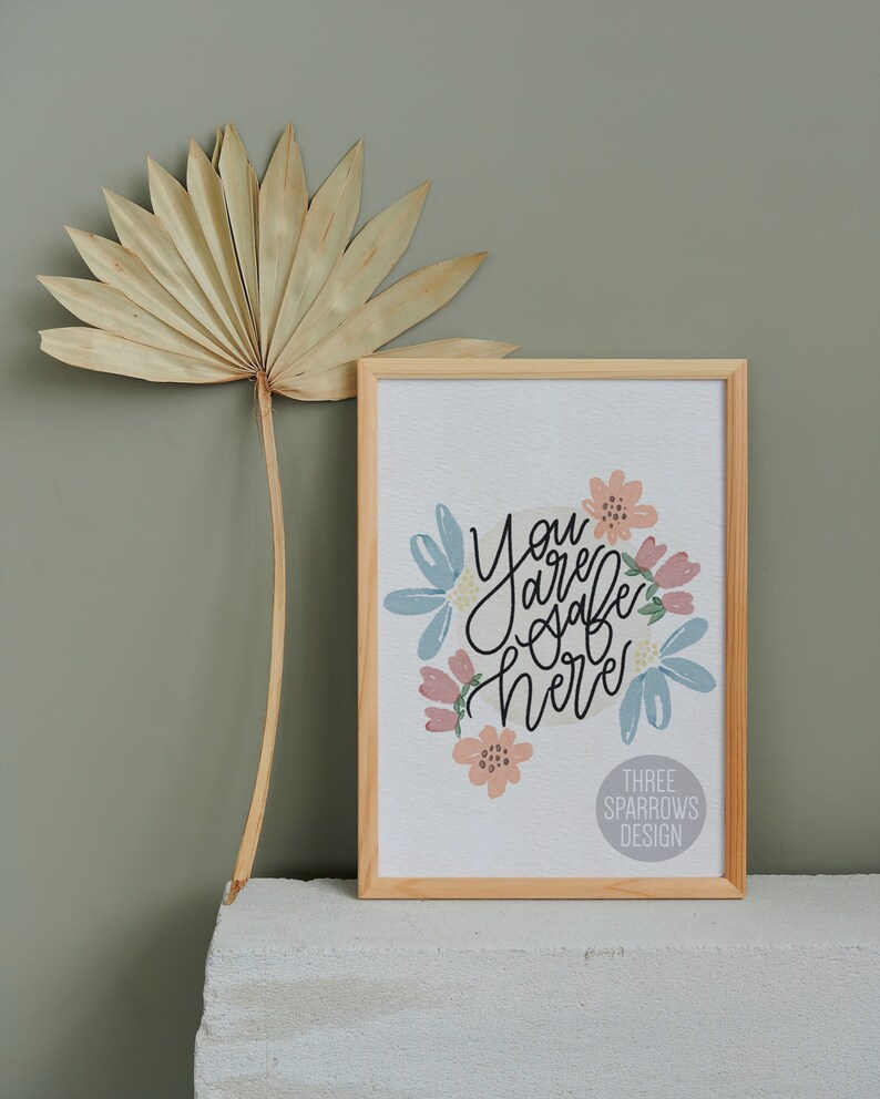 May include: A framed print with the words "You are safe here" in black script, surrounded by pastel-colored floral illustrations. The frame is light wood. A dried palm leaf is to the left.