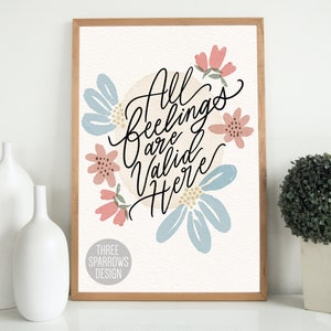 May include: A framed art print with the words "All Feelings Are Valid Here" in black script, surrounded by light blue and pink floral illustrations. The print has a light beige background and a wooden frame. The Three Sparrows Design logo is at the bottom.