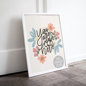 May include: A framed art print with the words "You are safe here" in black script, surrounded by watercolor flowers in shades of blue, pink, and peach. The print is in a white frame and features the text "THREE SPARROWS DESIGN".