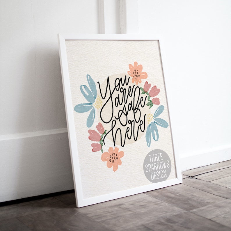 May include: A framed art print with the words "You are safe here" in black script, surrounded by watercolor flowers in shades of blue, pink, and peach. The print is in a white frame and features the text "THREE SPARROWS DESIGN".