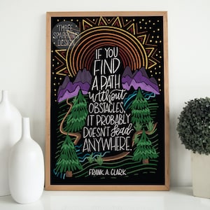 May include: A framed art print with a black background features colorful illustrations and the quote, "If you find a path without obstacles, it probably doesn't lead anywhere." The artwork includes mountains, trees, and a sun design. The artist's name, Frank A. Clark, is also included.