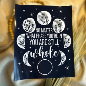 May include: A dark blue print with silver lettering and illustrations of moons and flowers. The text reads "No matter what phase you're in you are still whole".