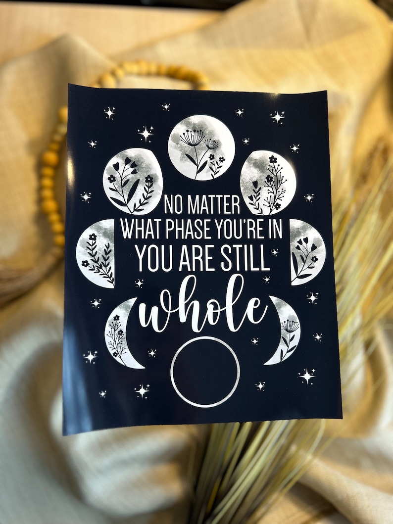 May include: A dark blue print with silver lettering and illustrations of moons and flowers. The text reads "No matter what phase you're in you are still whole".
