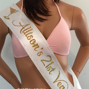 K&ouml;nnte beinhalten: Wei&szlig;er Satinschleife mit goldener Glitzerbeschriftung "Alison's 21st Vegas!" mit Strassbesatz und zwei Martini-Glas-Symbolen. Goldene gl&auml;nzende Beschriftung, Strassbesatz, 2 BLING - Martini-Gl&auml;ser.