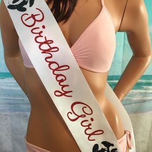 May include: White satin sash with red glitter text that reads "Birthday Girl". The sash has a black and white Mickey Mouse silhouette on each end.