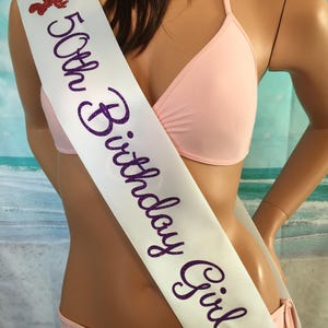 May include: White satin sash with purple glitter lettering that reads "50th Birthday Girl". The sash has two red glitter lobster designs on either end.