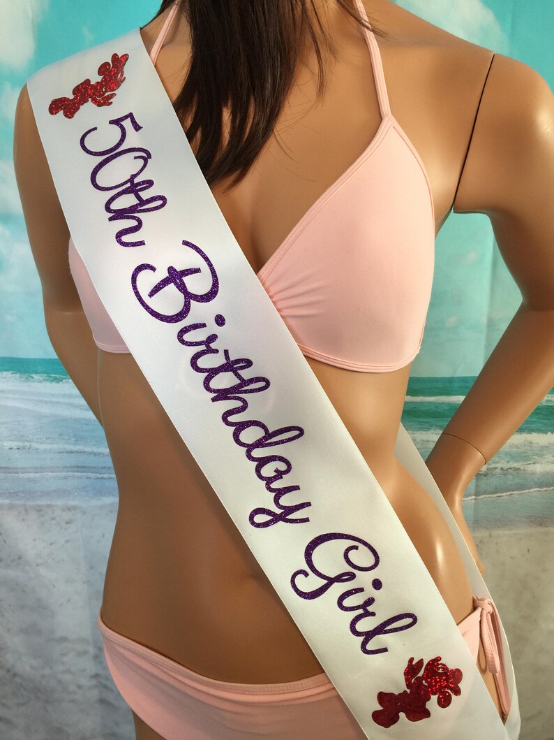 May include: White satin sash with purple glitter lettering that reads "50th Birthday Girl". The sash has two red glitter lobster designs on either end.