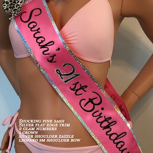 K&ouml;nnte beinhalten: Ein rosa Satinband mit silbernem Besatz und dem Text "Sarah's 21st Birthday". Das Band ist mit Strasssteinen und einer Schleife im Leopardenmuster verziert.