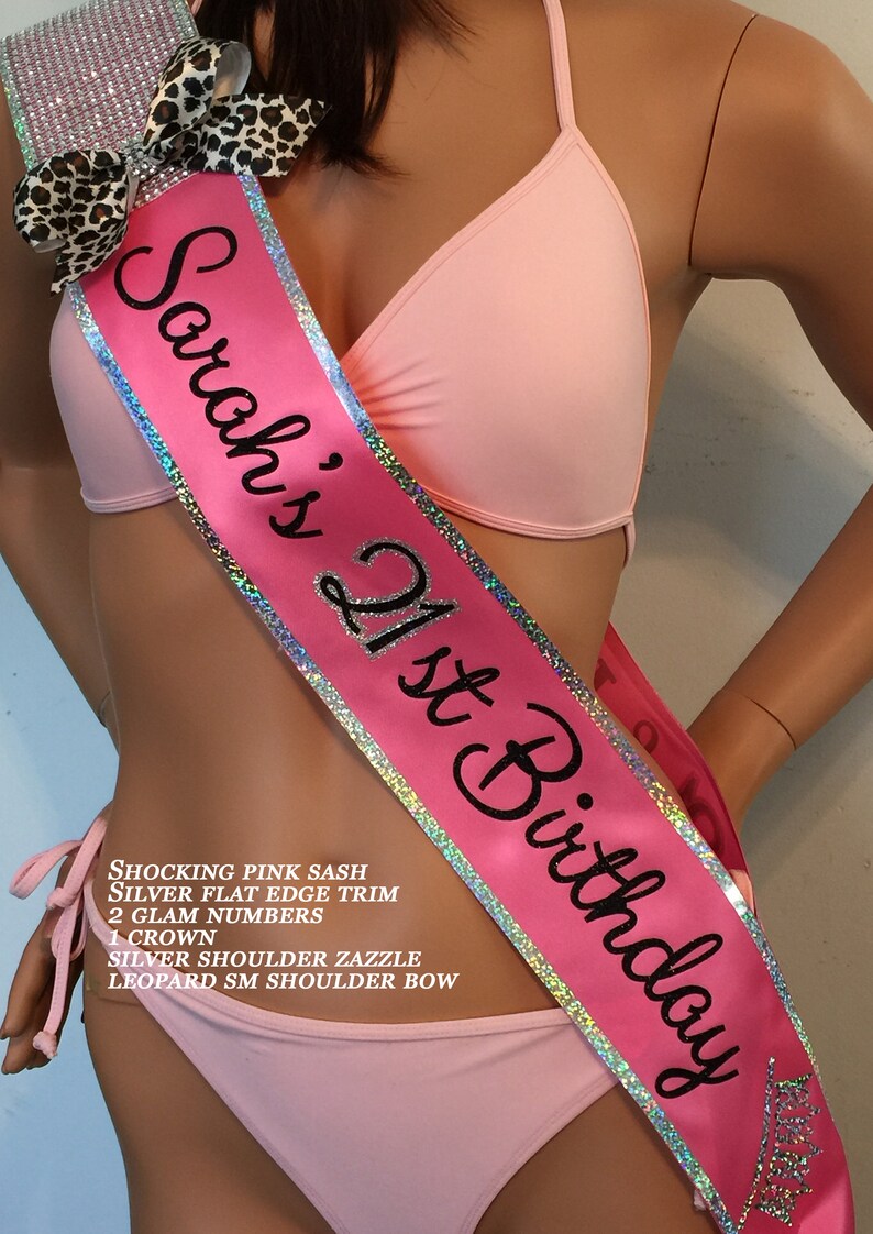 K&ouml;nnte beinhalten: Ein rosa Satinband mit silbernem Besatz und dem Text "Sarah's 21st Birthday". Das Band ist mit Strasssteinen und einer Schleife im Leopardenmuster verziert.